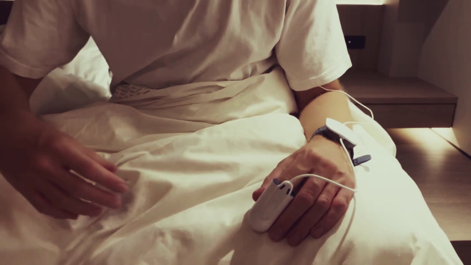 Iemand zit op de rand van het bed met de polsunit en de vingersensor van de thuis-slaaptest om. Warm lamplicht, gezicht buiten beeld.